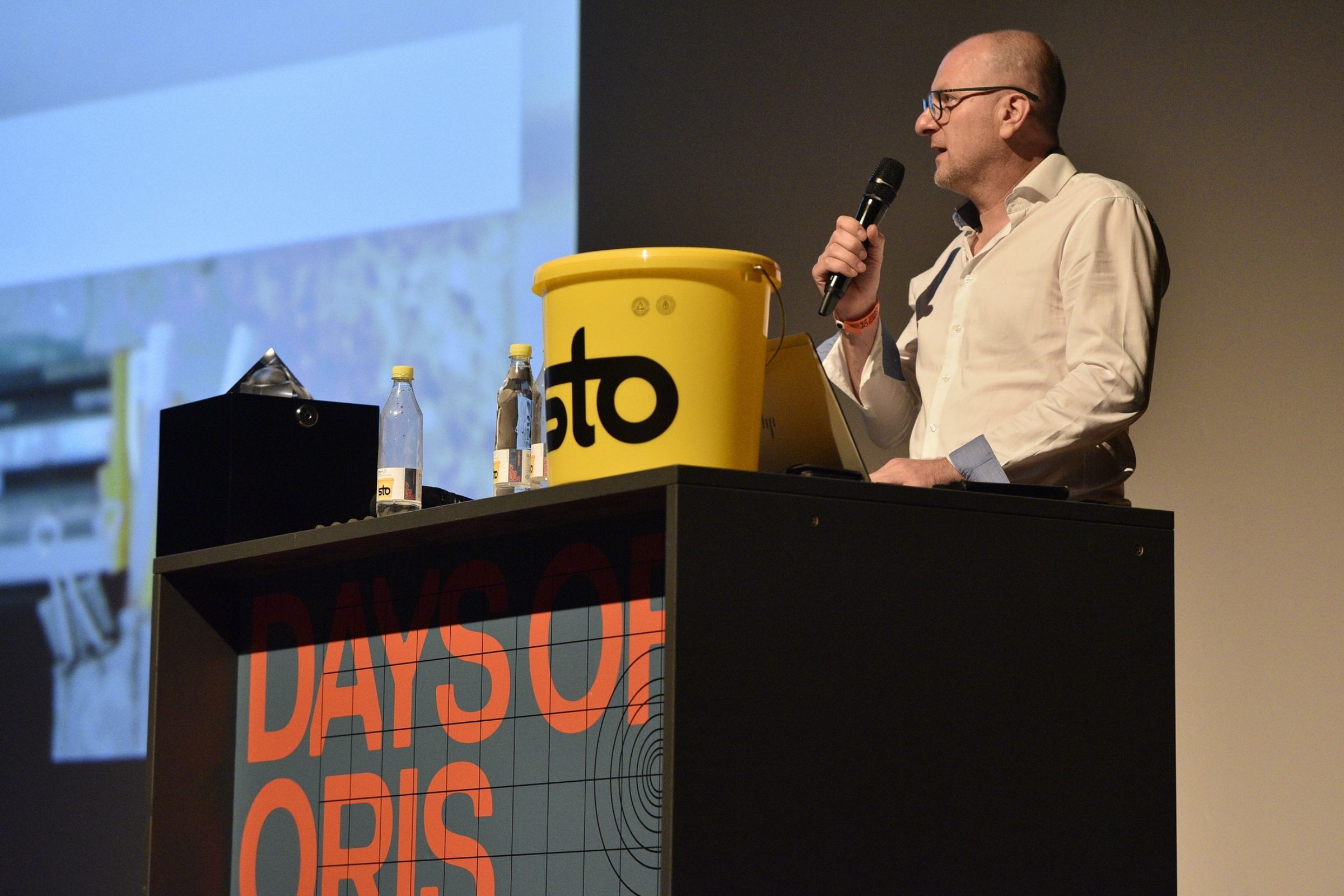 Govornik drži mikrofon iza podija „Days of Oris“ sa žutom Sto kantom.